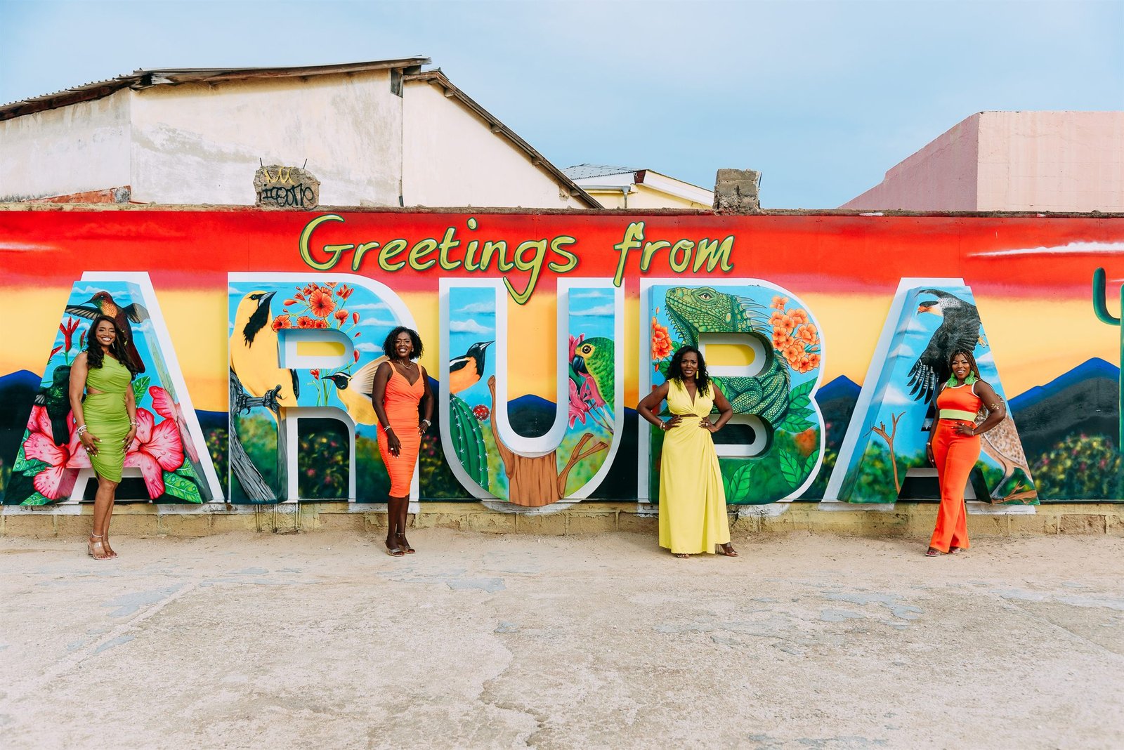 The four authors — Marla Lewis, Karen S. Wells, Kelly Copes-Anderson and Stacia Gowens — standing together in bright dresses before a vivid 'Greetings from Aruba' mural